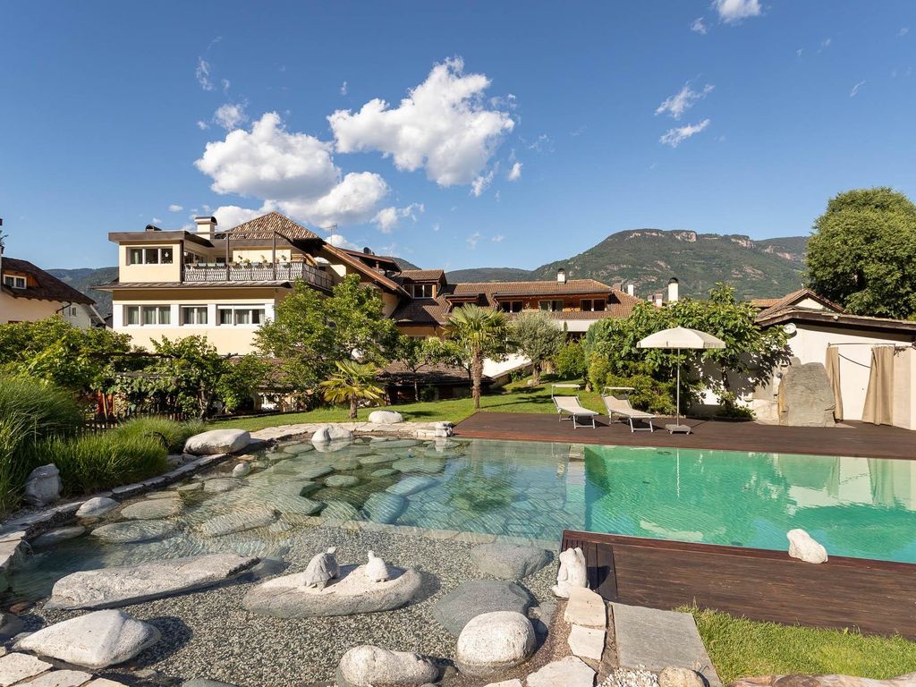 Im Vordergrund der Naturpool mit Teichbereich und Sonnenholzterrasse. Zwischen Pool und dem Gutshof Kreuzwegerhof Nals aus dem 16. Jahrhundert liegt eine grüne Liegewiese mit Palmen und verschiedenen Obstbäumen. Dahinter zeigt sich der Kreuzwegerhof Nals