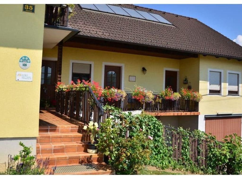 Die Außenansicht des Bauernhofs mit gelber Fassade, einem Balkon mit Blumenkästen und einer Eingangstreppe, ergänzt durch Solarpaneele auf dem Dach.