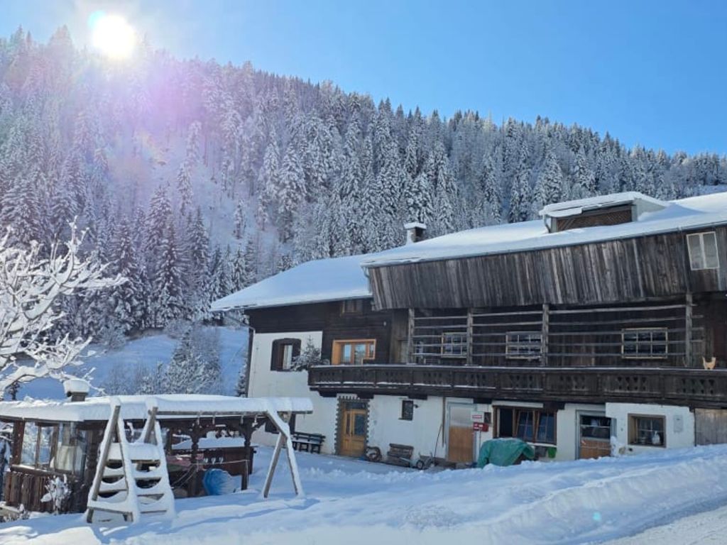 Der Bauernhof in einer verschneiten Winterlandschaft mit einem Wald im Hintergrund und einem Spielgerüst im Vordergrund.