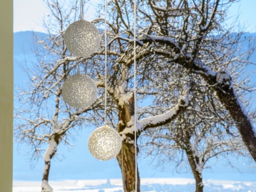 Blick aus dem Fenster des Bauernhofs auf eine verschneite Winterlandschaft mit Bäumen und dekorativen Kugeln im Vordergrund.