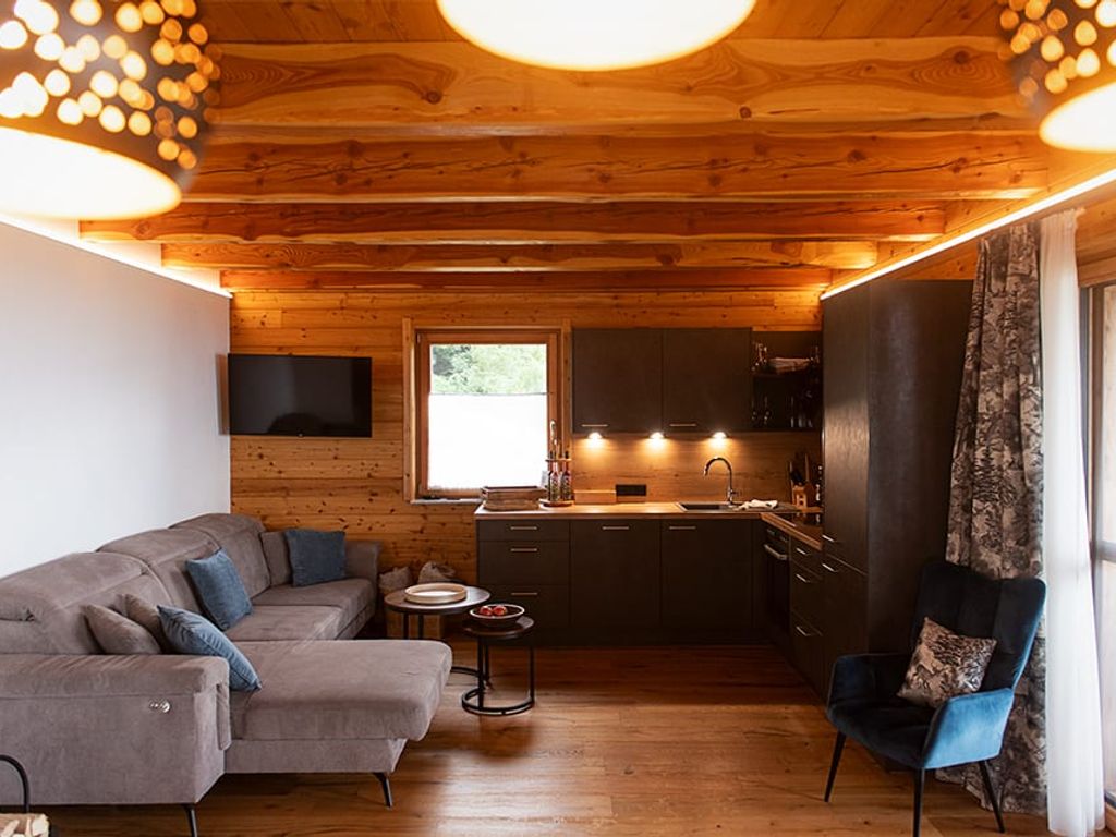 Wohnbereich des Ferienhauses mit Sofa, Fernseher, integrierter Küche und Holzbalkendecke.