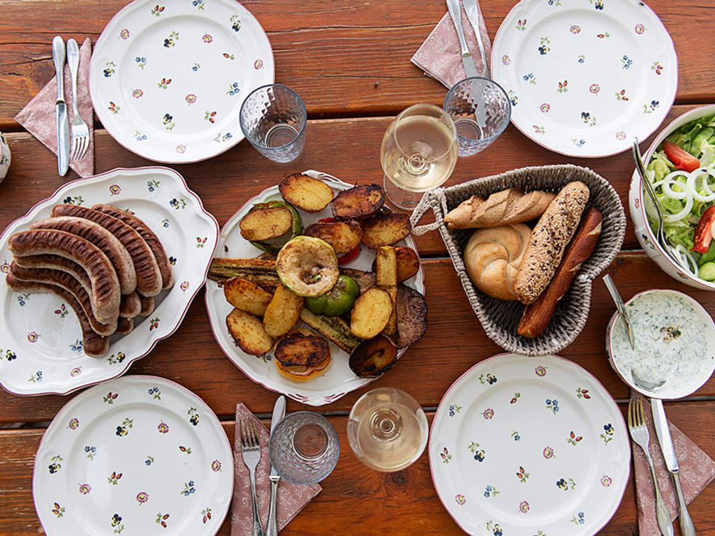 Ein gedeckter Tisch im Chalet mit gegrillten Würstchen, Ofengemüse, frischem Salat und Brot, ideal für eine Mahlzeit im Freien.