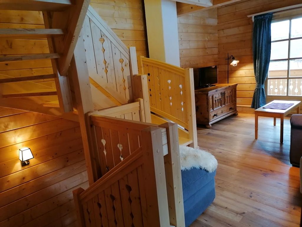 Der Innenbereich des Hofes präsentiert eine Holztreppe mit geschnitzten Geländerdetails, Holzwände und -böden sowie einen Wohnbereich mit Fernseher, Holztruhe und Fenster.