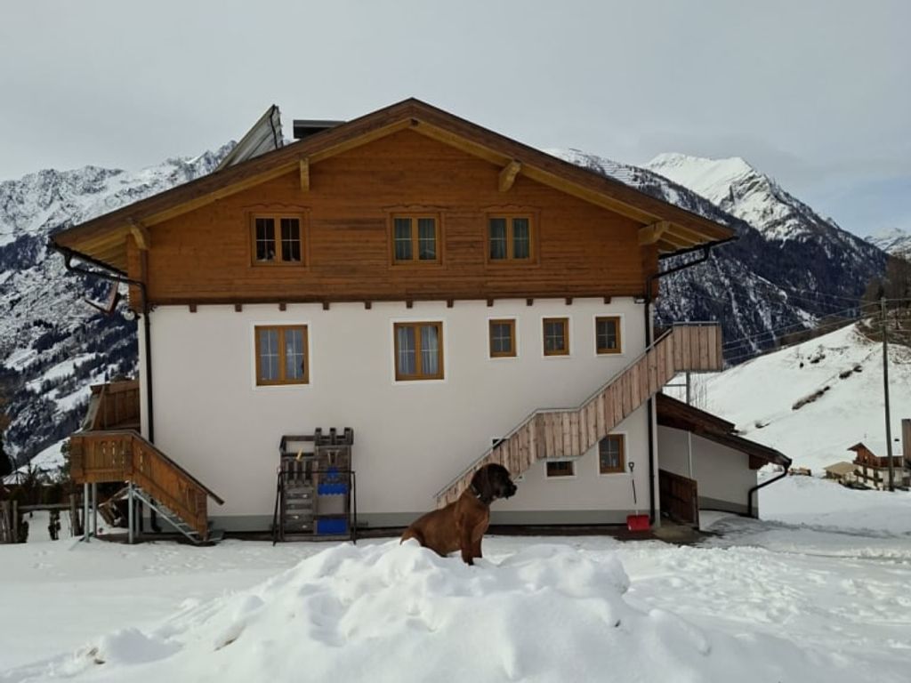 Außenansicht des Bauernhofs im Winter mit Schnee, Bergen im Hintergrund und einem Hund im Vordergrund.