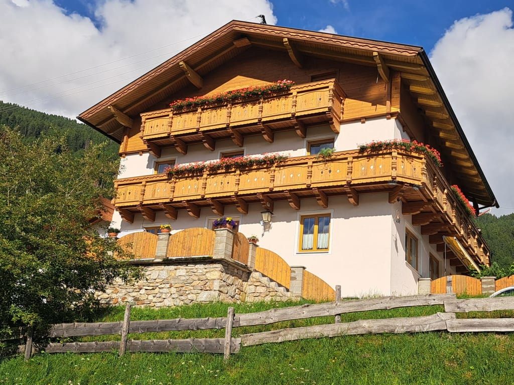 Der Bauernhof mit traditioneller Holzarchitektur, mehreren Blumenbalkonen und einer umliegenden Wiese mit Holzzaun.