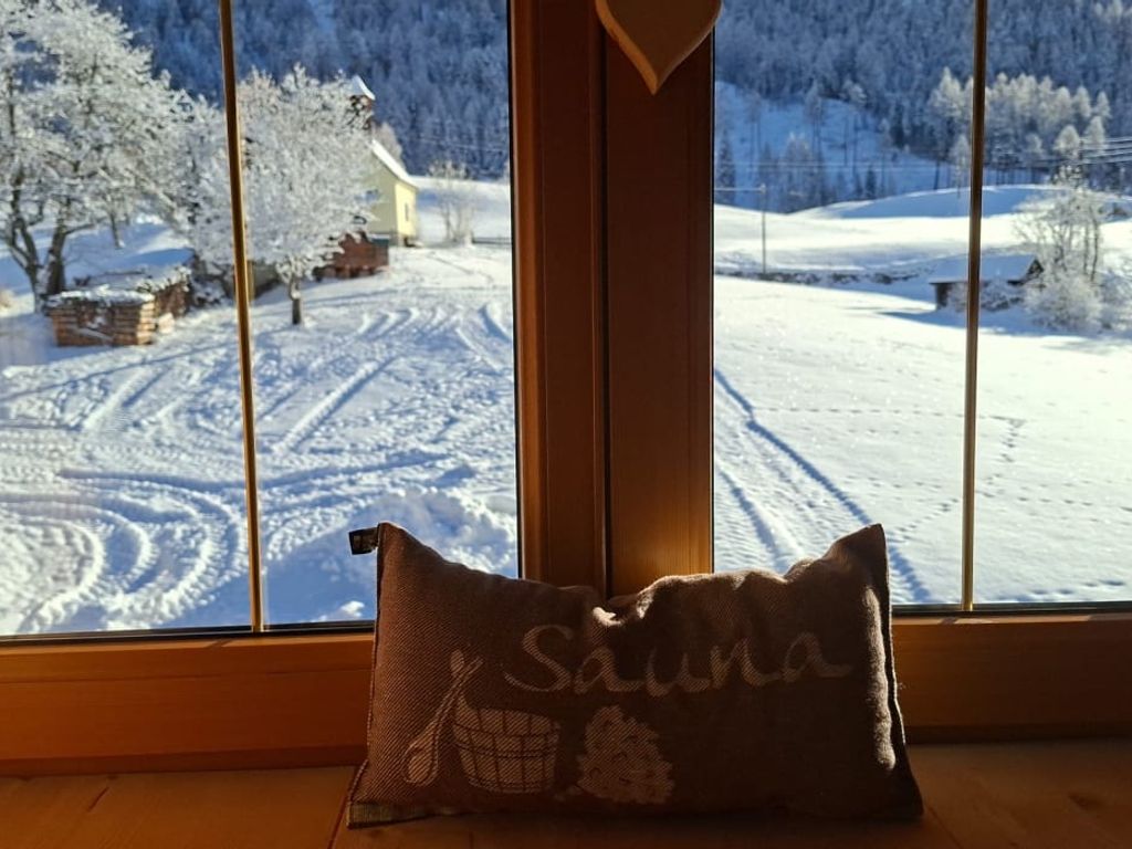 Blick aus dem Fenster des Bauernhofs auf die verschneite Winterlandschaft, mit einem Saunakissen auf der Fensterbank.