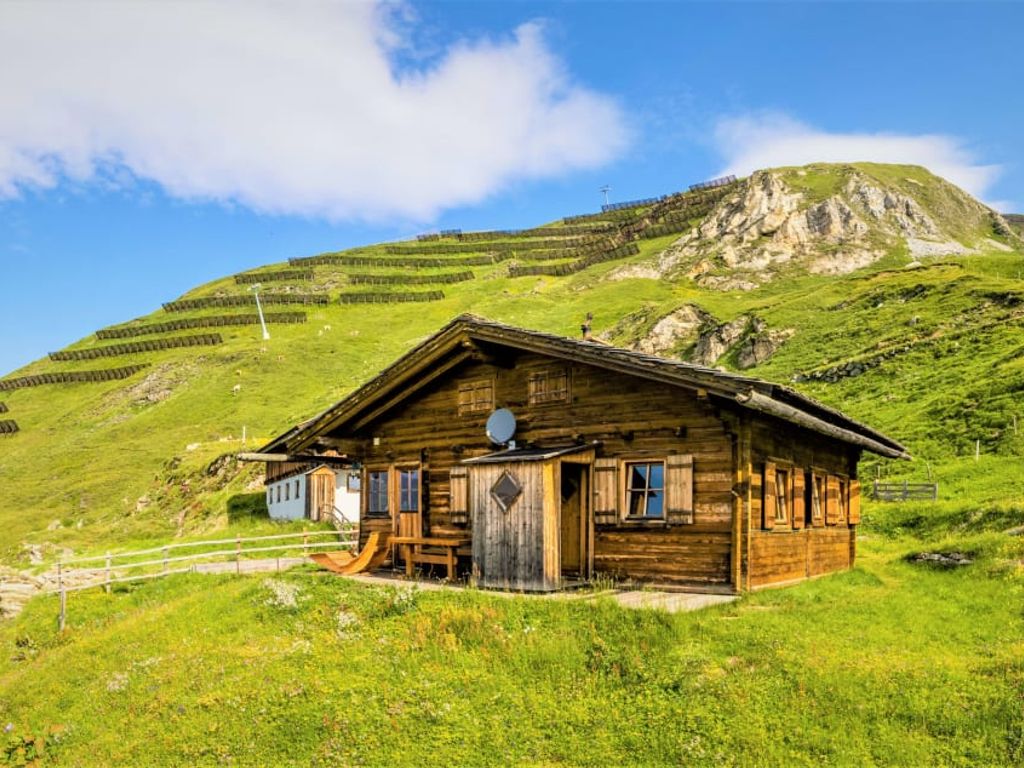 Die Almhütte in Holzbauweise liegt auf einem grünen Berghang und bietet einen Sitzbereich im Freien.