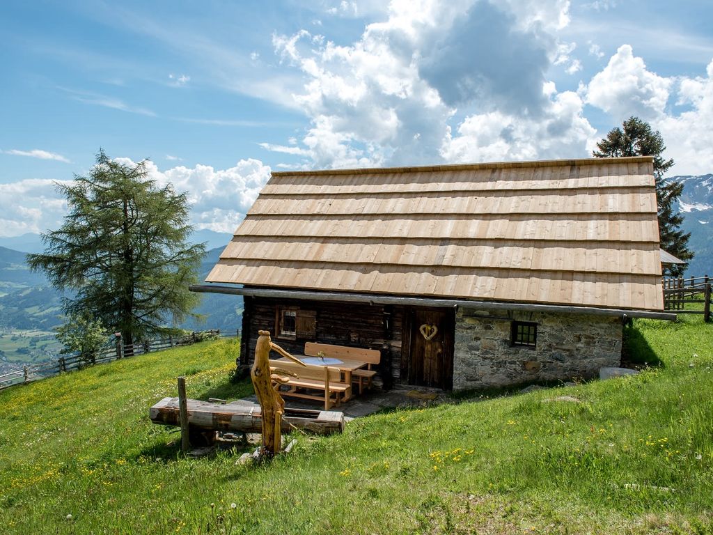 Die Alm mit Holzdach, Außenbereich mit Holztisch und Bänken sowie einem hölzernen Wasserbecken, gelegen an einem Hang mit Bergblick.