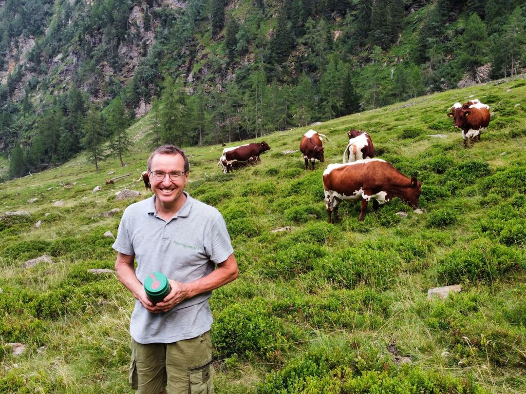 Der Gastgeber des Bauernhofs auf einer Almwiese mit weidenden Kühen und der umgebenden Berglandschaft.