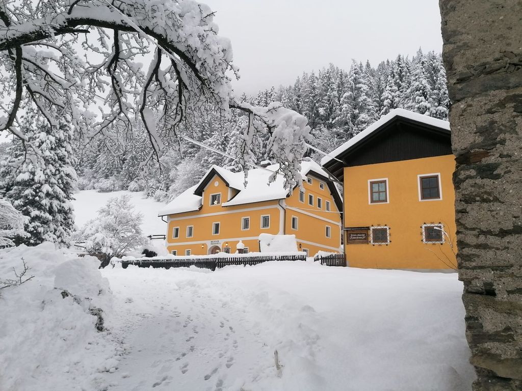 Der gelbe Bauernhof mit schneebedecktem Dach, umgeben von verschneiten Bäumen in der Winterlandschaft.