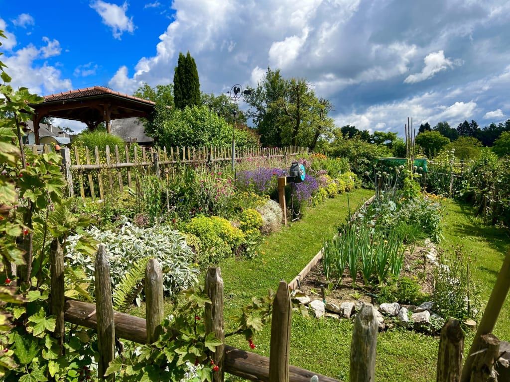 Der Garten des Bauernhofs mit Holzzaun, vielfältigen Blumenbeeten und einem Gemüseanbau.