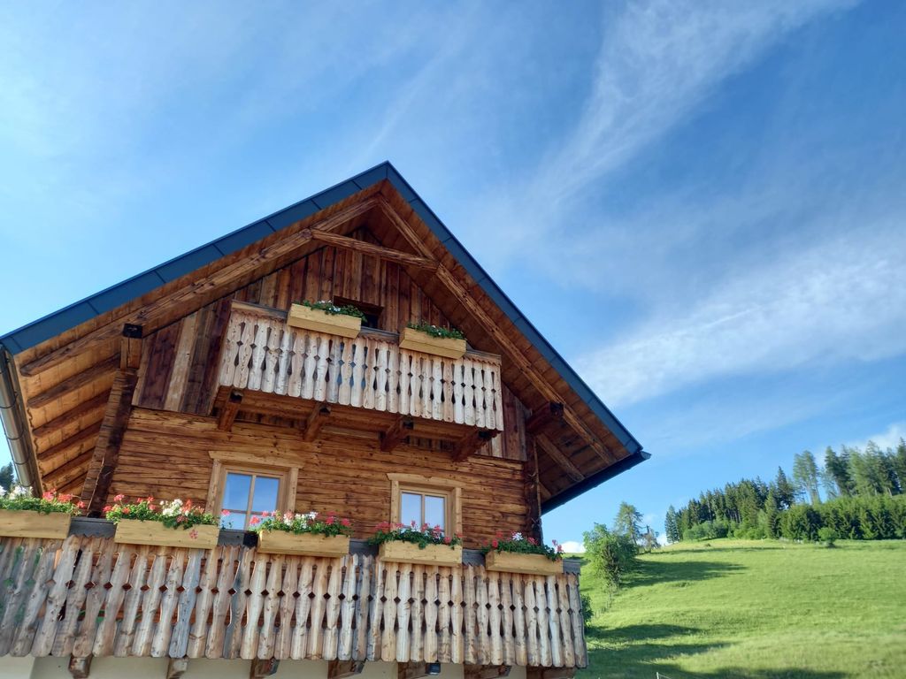 Aussenansicht der Alm aus Holz mit zwei Balkonen und Blumenkästen vor einer grünen Wiese.