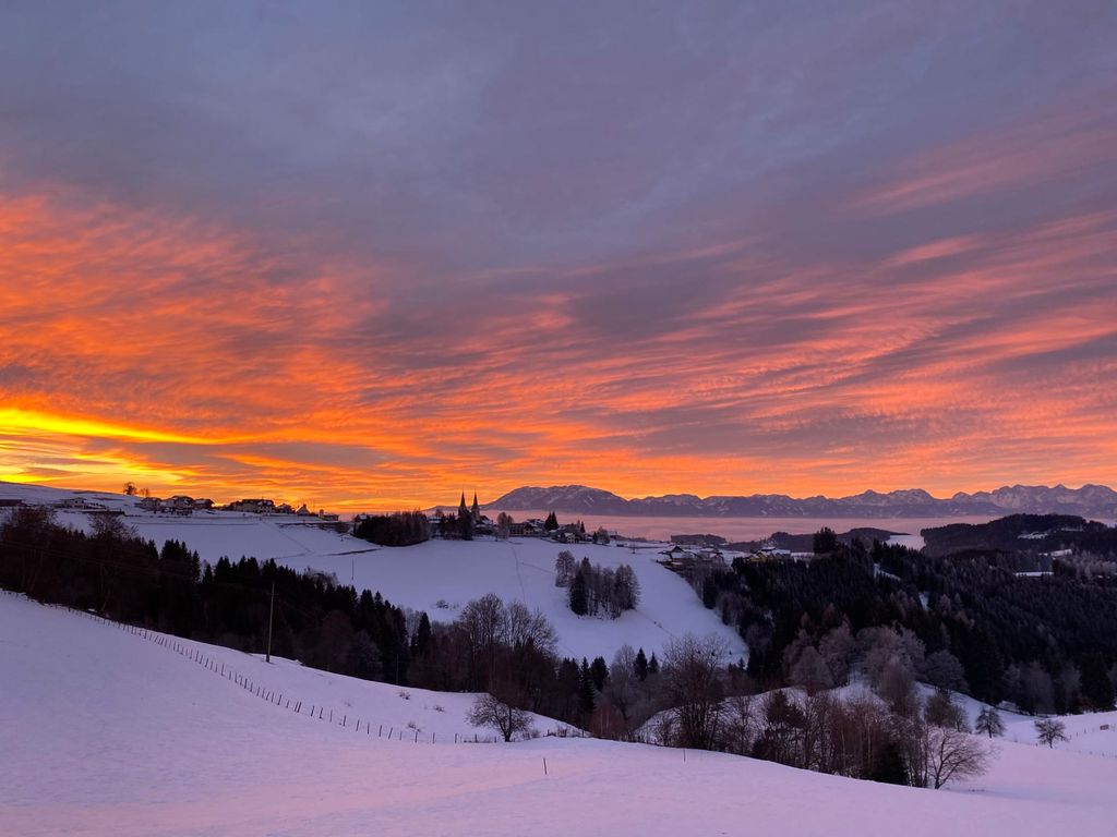 Blick auf die winterliche Landschaft mit dem Dorf, den Bergen und einem orange-lila Himmel vom Bauernhof aus.