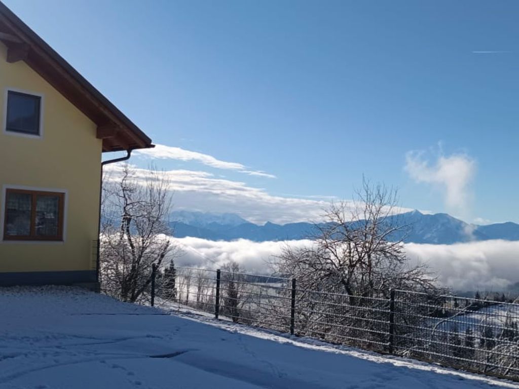Der gelbe Bauernhof mit Garage und einer schneebedeckten Auffahrt, mit Blick auf ein Bergpanorama über einer Wolkendecke.