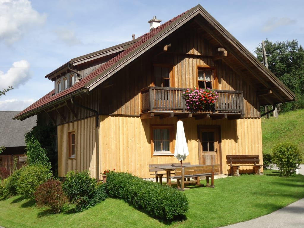 Die Alm von außen mit Balkon, blühenden Blumenkästen und einer Sitzgruppe mit Sonnenschirm auf der Rasenfläche.