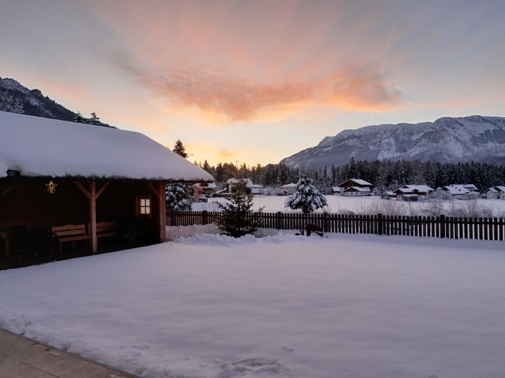 Winterlandschaft des Bauernhofs mit schneebedecktem Garten, Holzpavillon und Bergen im Hintergrund.