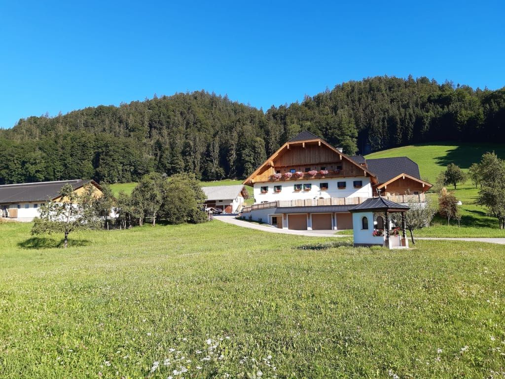 Der Bauernhof mit blumengeschmückten Balkonen, umgeben von grünen Wiesen und einem Wald, sowie einer kleinen Kapelle.