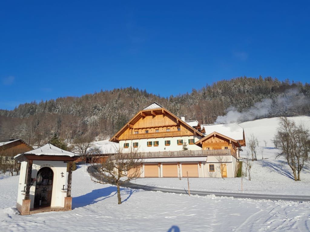 Der Bauernhof im Winter mit schneebedeckter Landschaft, Wald im Hintergrund und einem kleinen Schrein im Vordergrund.