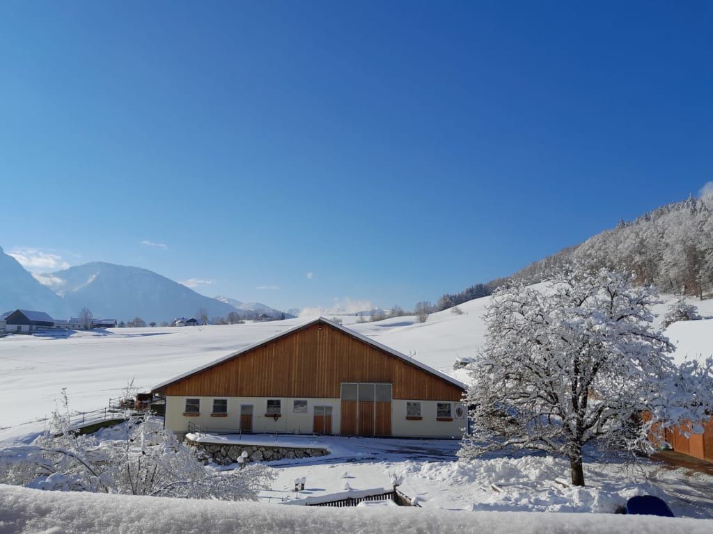 Der Bauernhof in einer verschneiten Winterlandschaft mit Bergen und blauem Himmel.