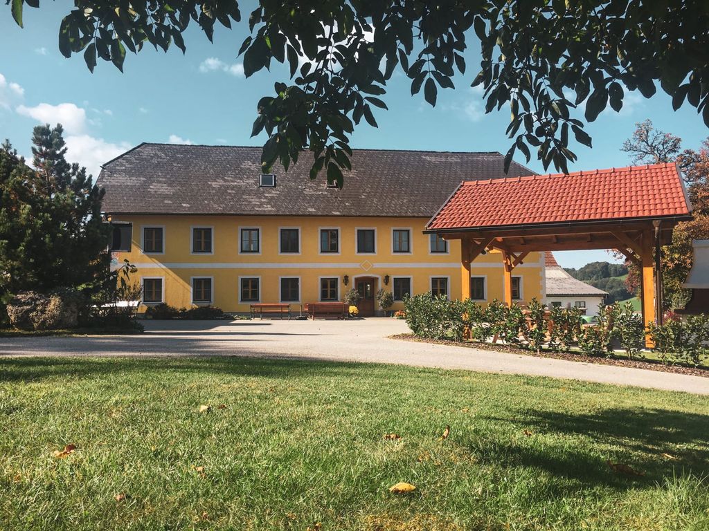 Das gelbe Bauernhofgebäude mit dunklem Dach, einem überdachten Holztor und Sitzbänken im Außenbereich, umgeben von Grünflächen.