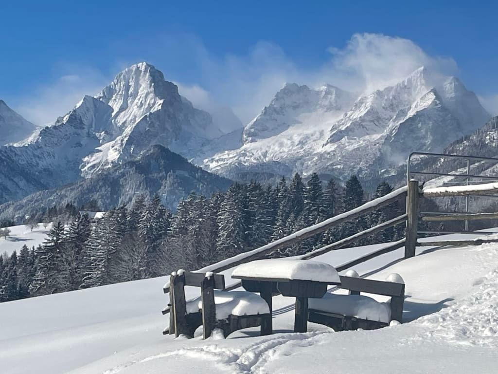 Blick auf die verschneiten Berge vom Bauernhof, mit einem schneebedeckten Holztisch und Bänken im Vordergrund.