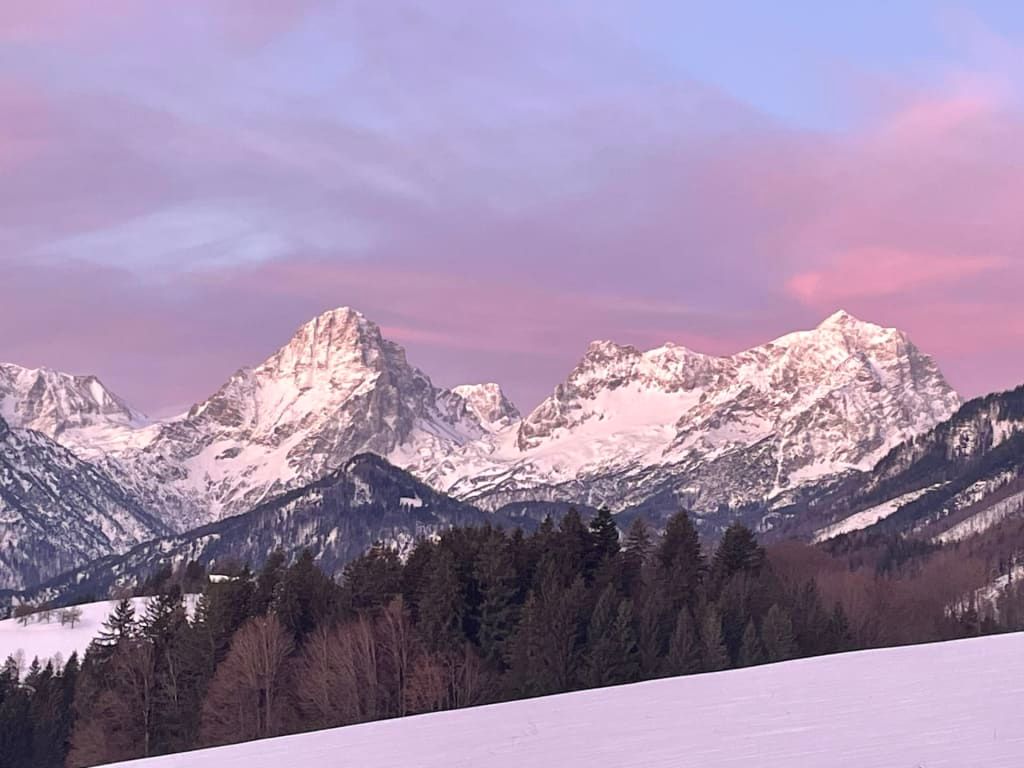 Blick auf schneebedeckte Berge und Wälder bei Sonnenuntergang vom Bauernhof aus.