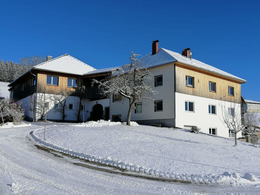 Außenansicht des Bauernhofs im Winter, mit schneebedeckten Dächern und einer Zufahrt unter klarem blauem Himmel.