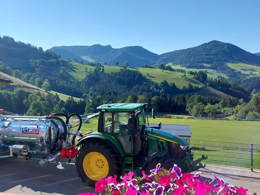 Blick vom Bauernhof auf die bergige Landschaft mit einem Traktor und blühenden Blumen im Vordergrund.