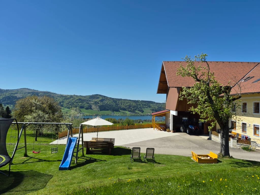 Außenansicht des Bauernhofs mit Garten, Kinderspielplatz, Sitzgelegenheiten und Blick auf den See und die Berge.