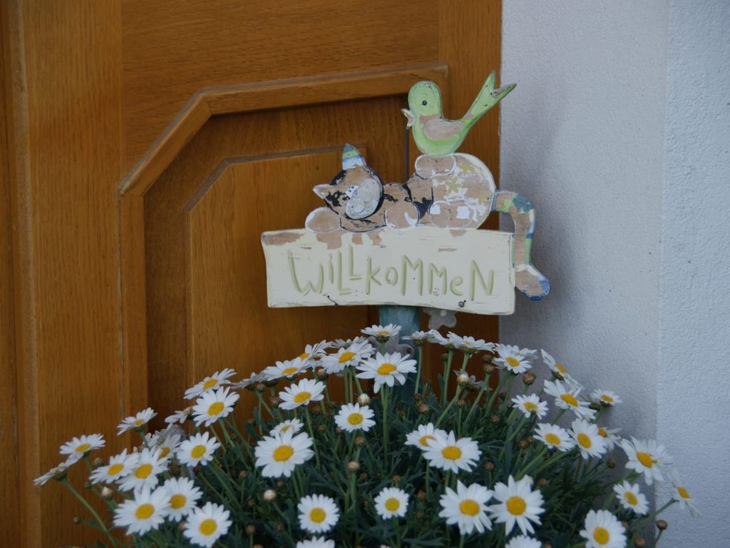 Das Willkommen-Schild mit dekorativer Katze und Vogel, neben einem Topf mit Gänseblümchen, empfängt Gäste am Bauernhof.