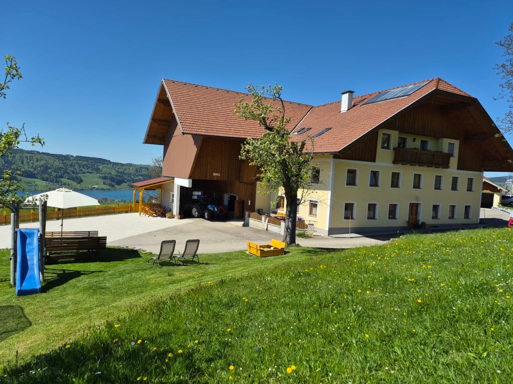 Der Bauernhof mit Gartenbereich, Rutsche, Sonnenliegen und überdachtem Sitzbereich, mit Blick auf den See und die Berge.