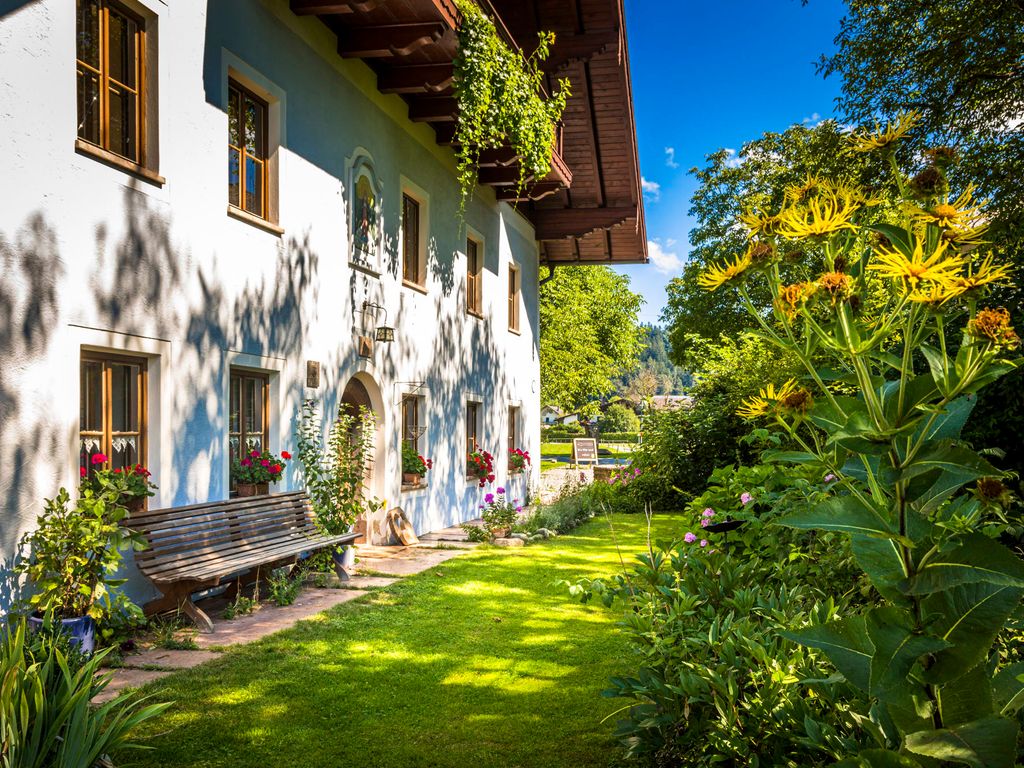 Der Außenbereich des Bauernhofs mit grüner Rasenfläche, blühenden Blumen und einer Holzbank vor dem Gebäude.