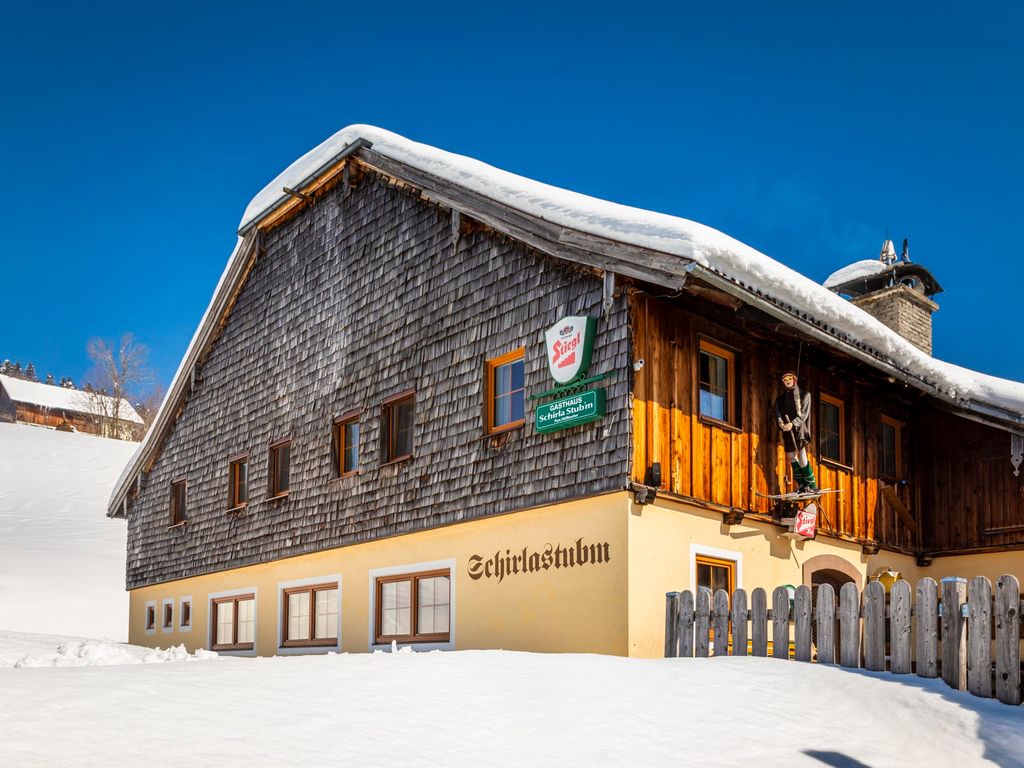 Der Bauernhof im Winter, mit einer Fassade aus Holzschindeln und gelbem Putz, sowie dem Gasthaus Scharla Stubn und verschneiter Umgebung.