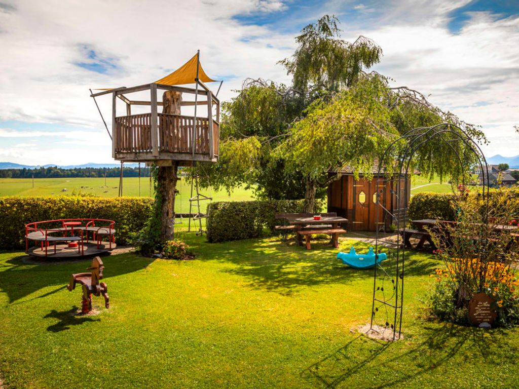 Der einladende Spielplatz des Bauernhofs mit einem hölzernen Baumhaus, verschiedenen Spielgeräten und gemütlichen Sitzbereichen im grünen Garten.