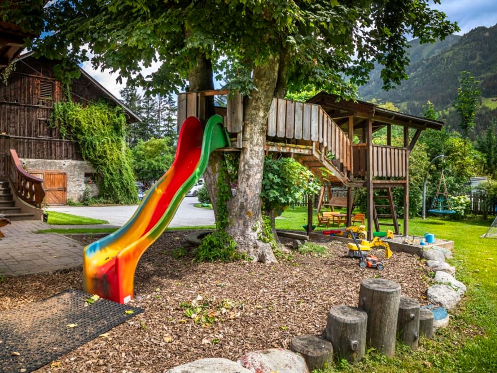 Der einladende Kinderspielplatz des Bauernhofs bietet eine bunte Rutsche, ein Baumhaus und eine Schaukel vor einer malerischen Bergkulisse.