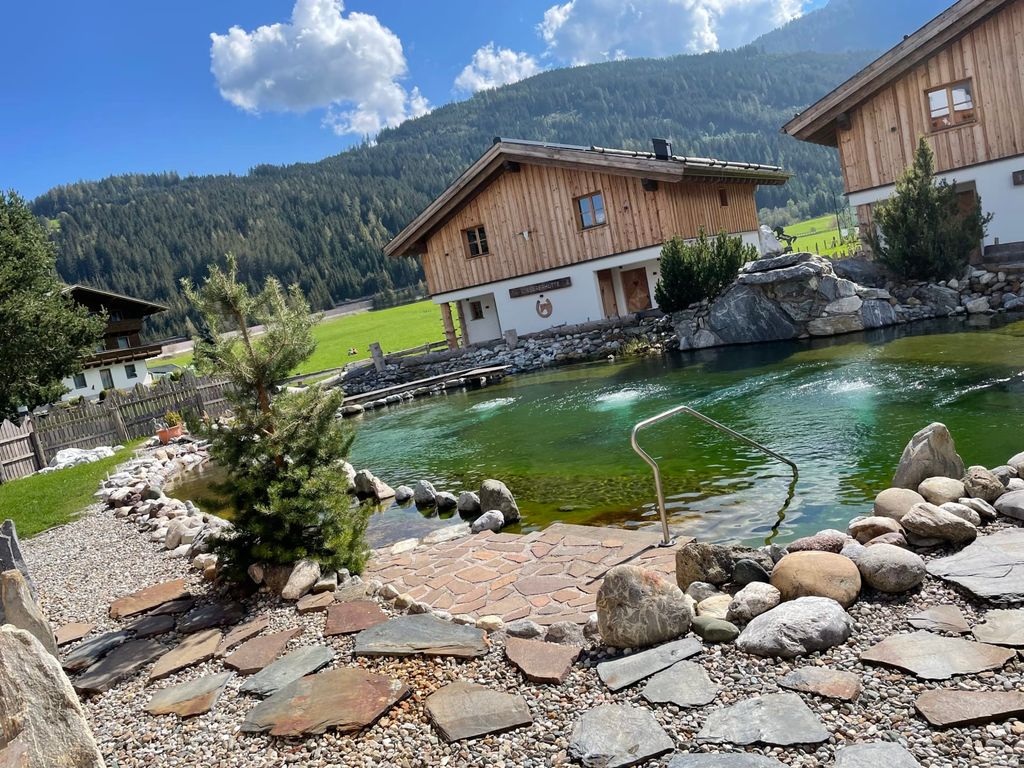 Naturbadeteich des Bauernhofs mit Einstiegsleiter und steinumrandetem Ufer vor einer Bergkulisse.