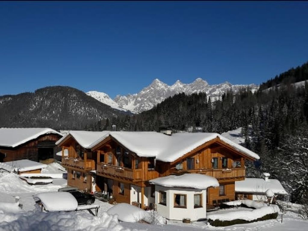 Der Bauernhof mit verschneiten Holzbalkonen und Dächern, umgeben von einer Winterlandschaft mit Blick auf die Alpen.