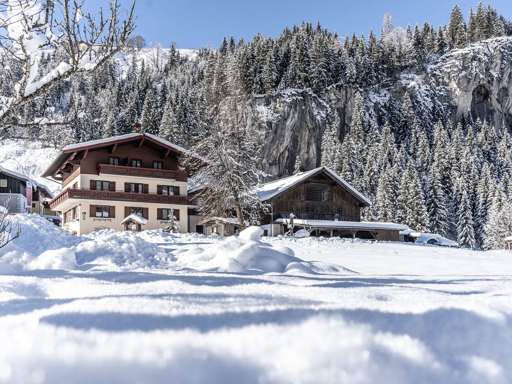 Der Bauernhof mit seinen Gebäuden in einer tief verschneiten Winterlandschaft vor Nadelwäldern und einer Felswand.