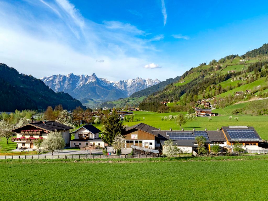 Der Bauernhof mit seinen Gebäuden liegt in einer grünen Tal-Landschaft, umgeben von Wiesen und Bergen, teilweise mit Solarpaneelen auf den Dächern.