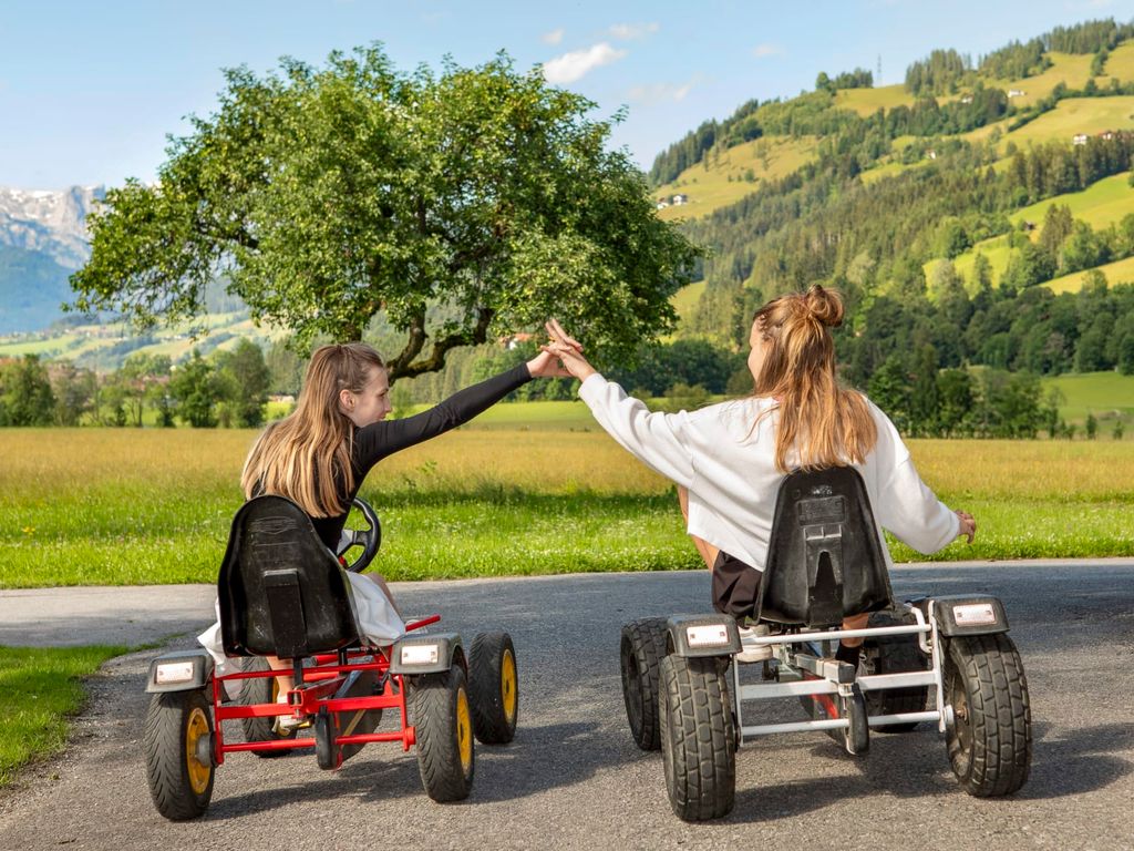 Zwei Kinder fahren auf dem Bauernhof mit Gokarts auf einem Weg vor der Kulisse der Berge.