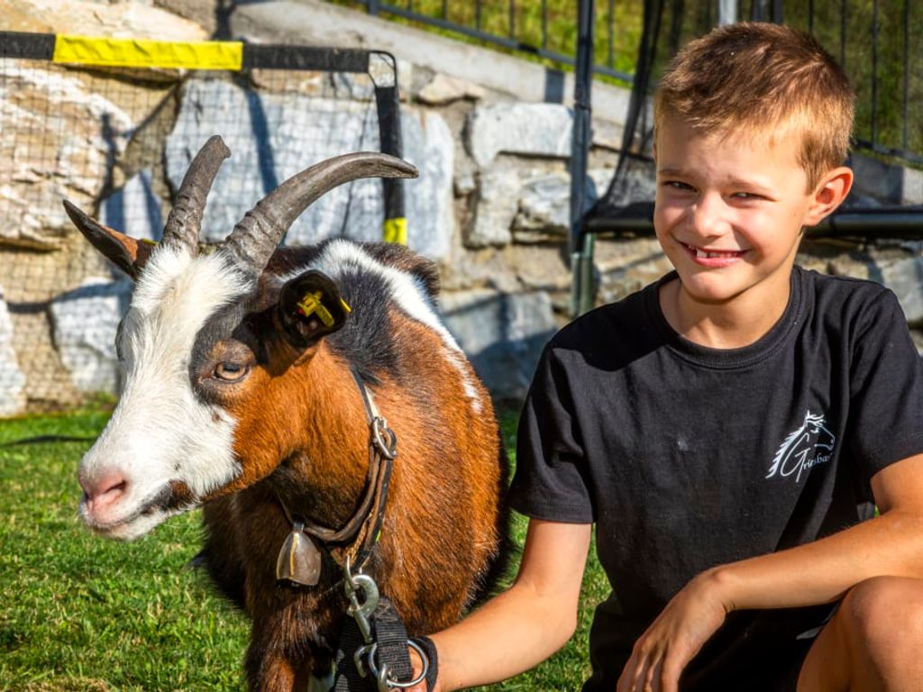 Ein Junge lächelt neben einer zahmen Ziege auf der Wiese des familienfreundlichen Bauernhofs.