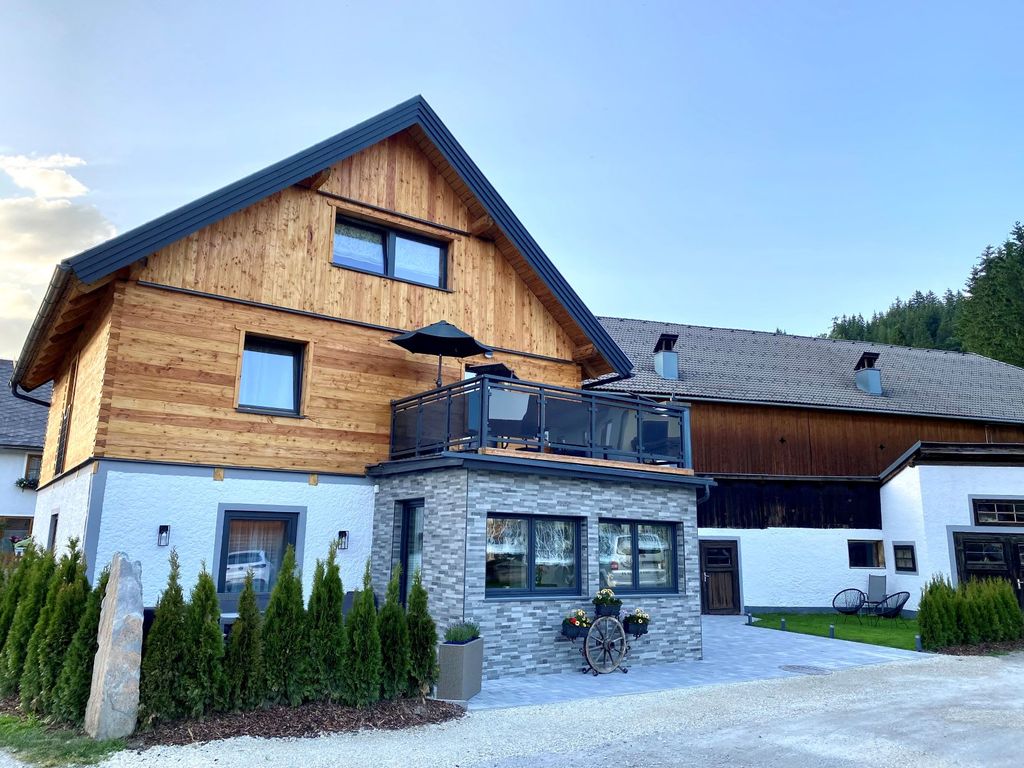 Die Außenansicht des Bauernhofs mit modernisierter Holzfassade, einem Balkon mit Sitzgelegenheiten und Sonnenschirm sowie dem angrenzenden Wirtschaftsgebäude.
