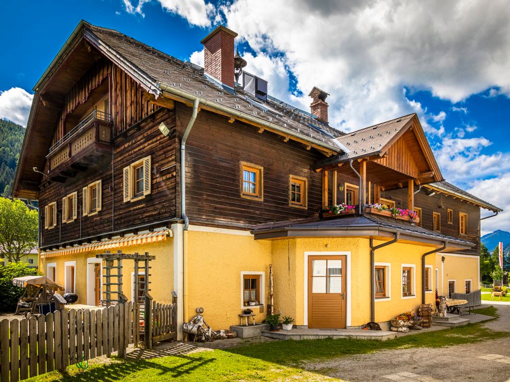 Der Bauernhof mit Holzfassade, gelb gestrichenem Erdgeschoss, mehreren Balkonen mit Blumen und einem umzäunten Gartenbereich.