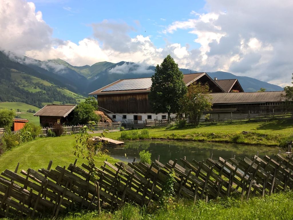 Der Bauernhof mit Teich, traditionellen Holzgebäuden und Blick auf die umliegende Berglandschaft.