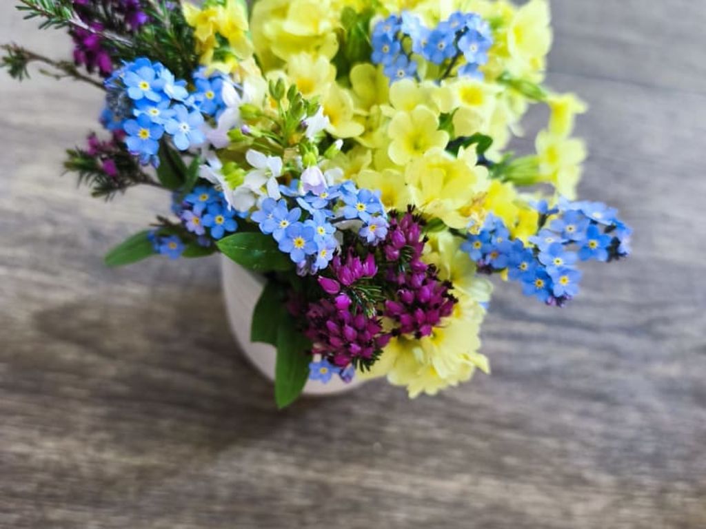 Ein kleiner Blumenstrauß mit gelben, blauen und violetten Blüten in einer weißen Vase auf einem Holztisch im Bauernhof.