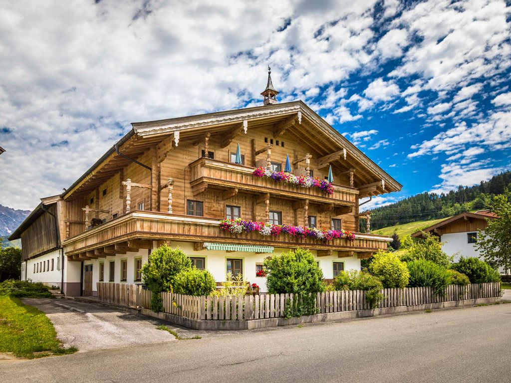 Außenansicht des Bauernhofs mit traditioneller Holzfassade, mehreren blumengeschmückten Balkonen und Sonnenschirmen.