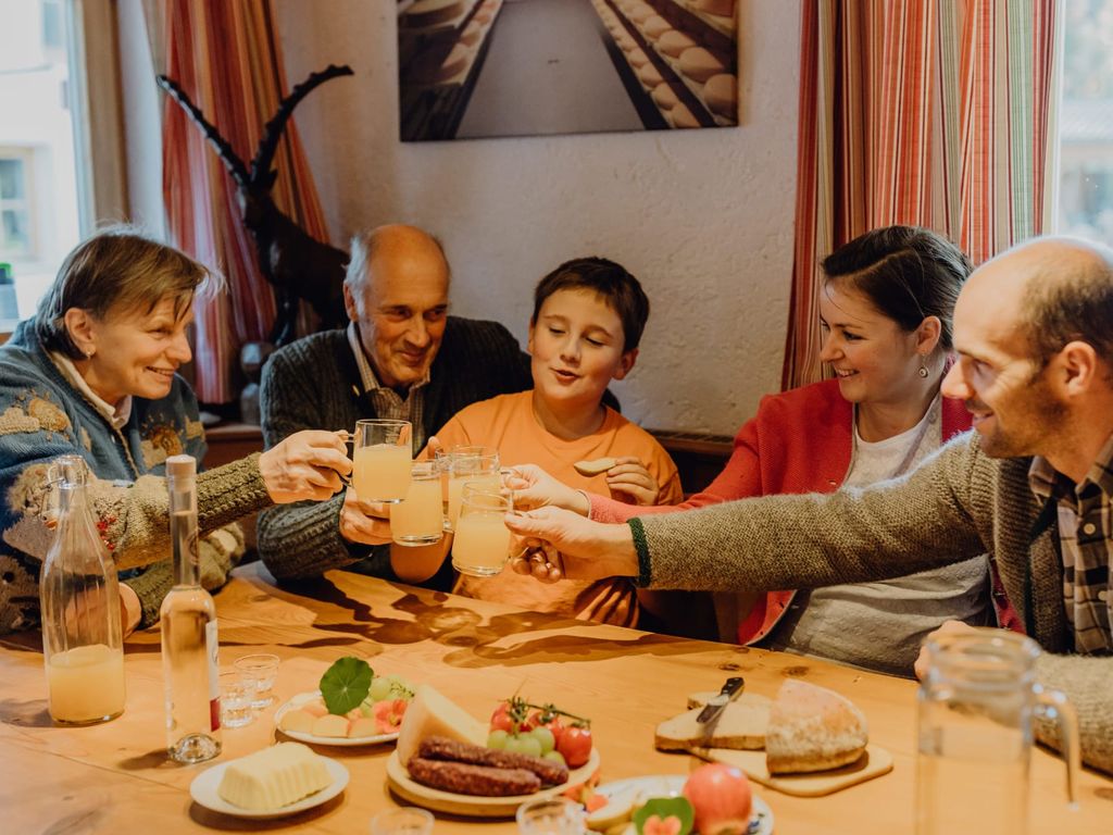 Familie bei einem Essen am Holztisch mit Käse, Wurst und hofeigenen Getränken auf dem Bauernhof.