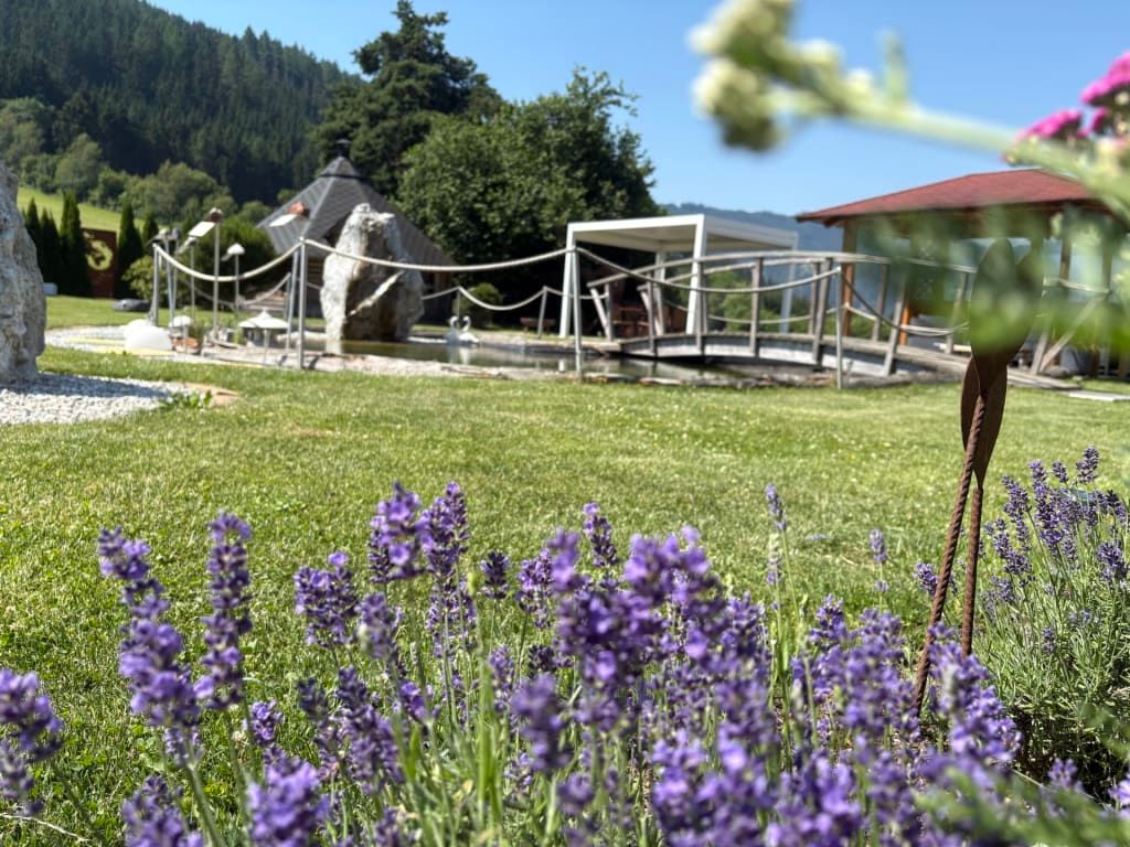 Die Gartenanlage des Landhofs mit Lavendel, einem Teich mit Brücke, einer Pergola und einem Pavillon, umgeben von Rasen und Bergen.