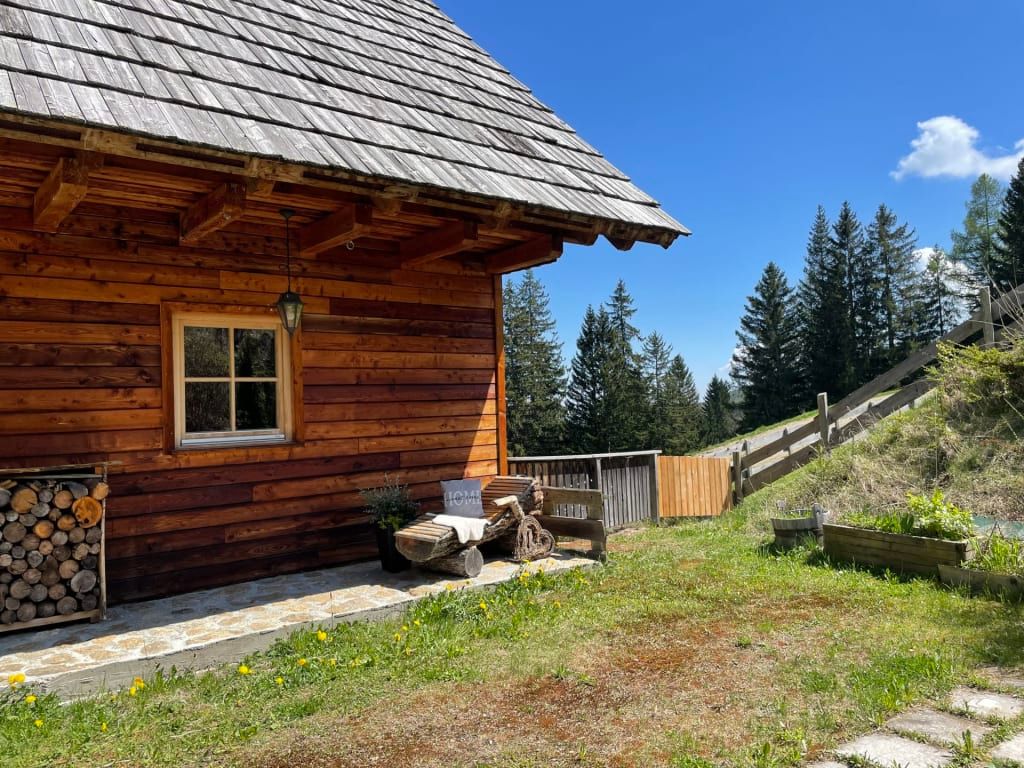 Außenansicht der Alm mit Holzfassade, Fenster, Holzstapel und einer Sitzbank aus Holzstämmen.
