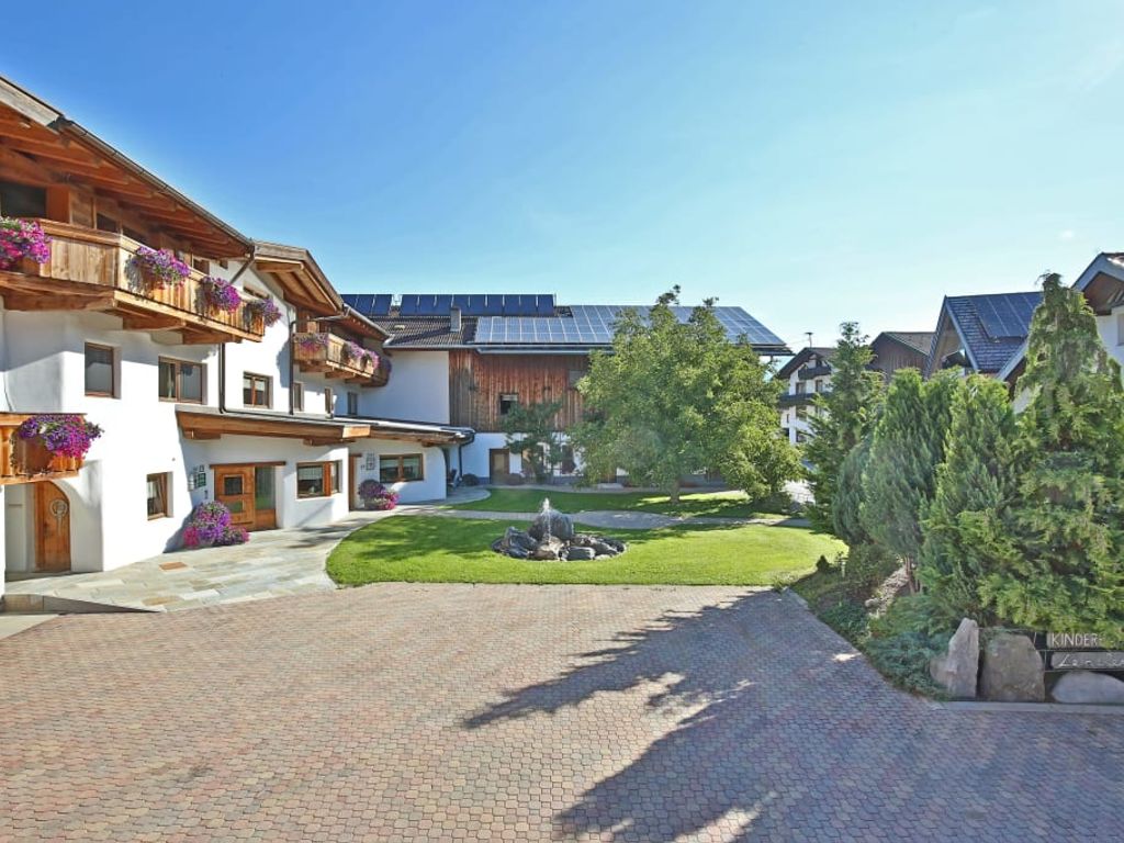 Außenansicht des Bauernhofs mit blumengeschmückten Holzbalkonen, gepflastertem Hof und einem Brunnen im Garten.